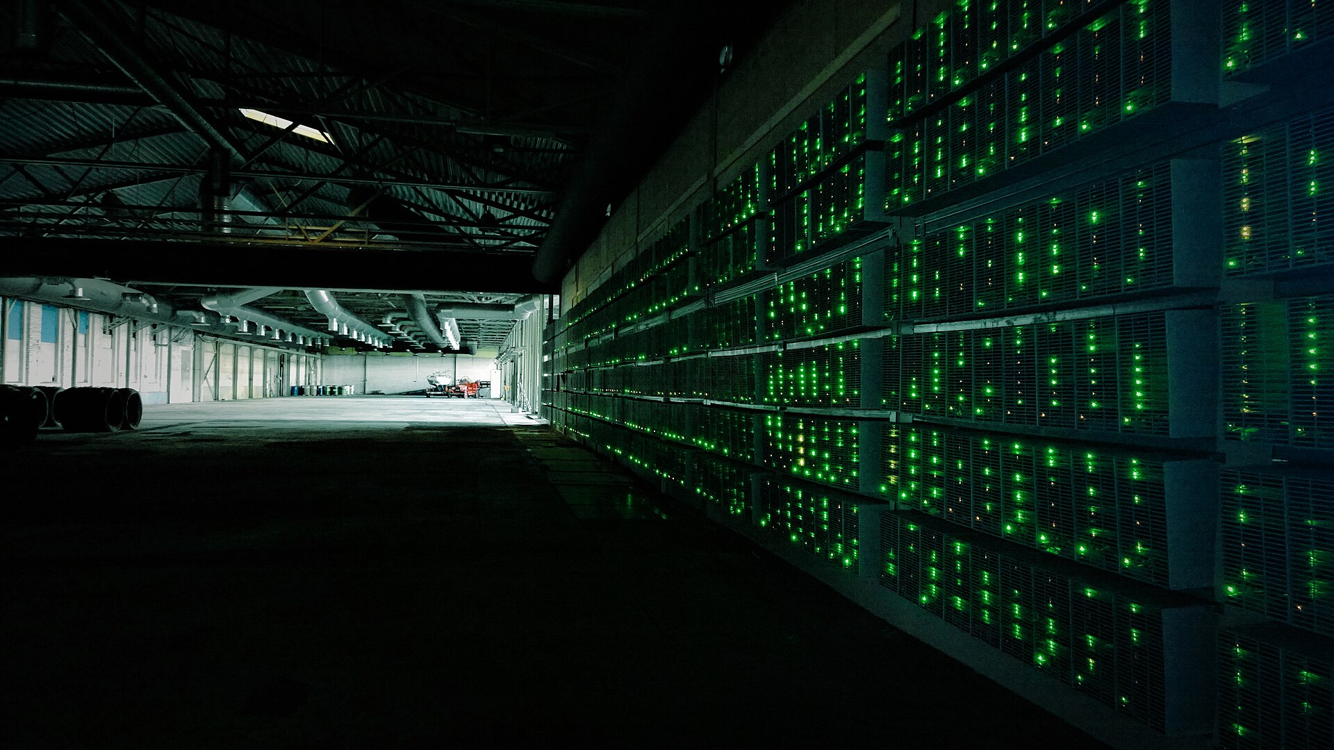 A dark industrial hall lined with rows of cryptocurrency mining machines and green status lights